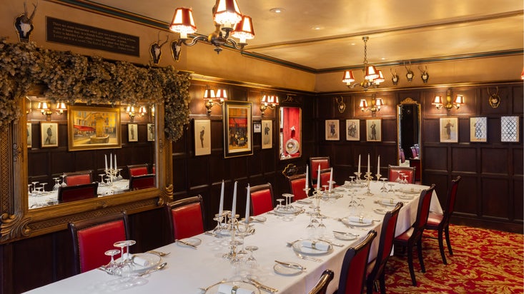 Elegant private dining room with dark wood-panelled walls, framed artwork, red leather chairs, white tablecloths, silver candlesticks, and warm chandelier lighting.